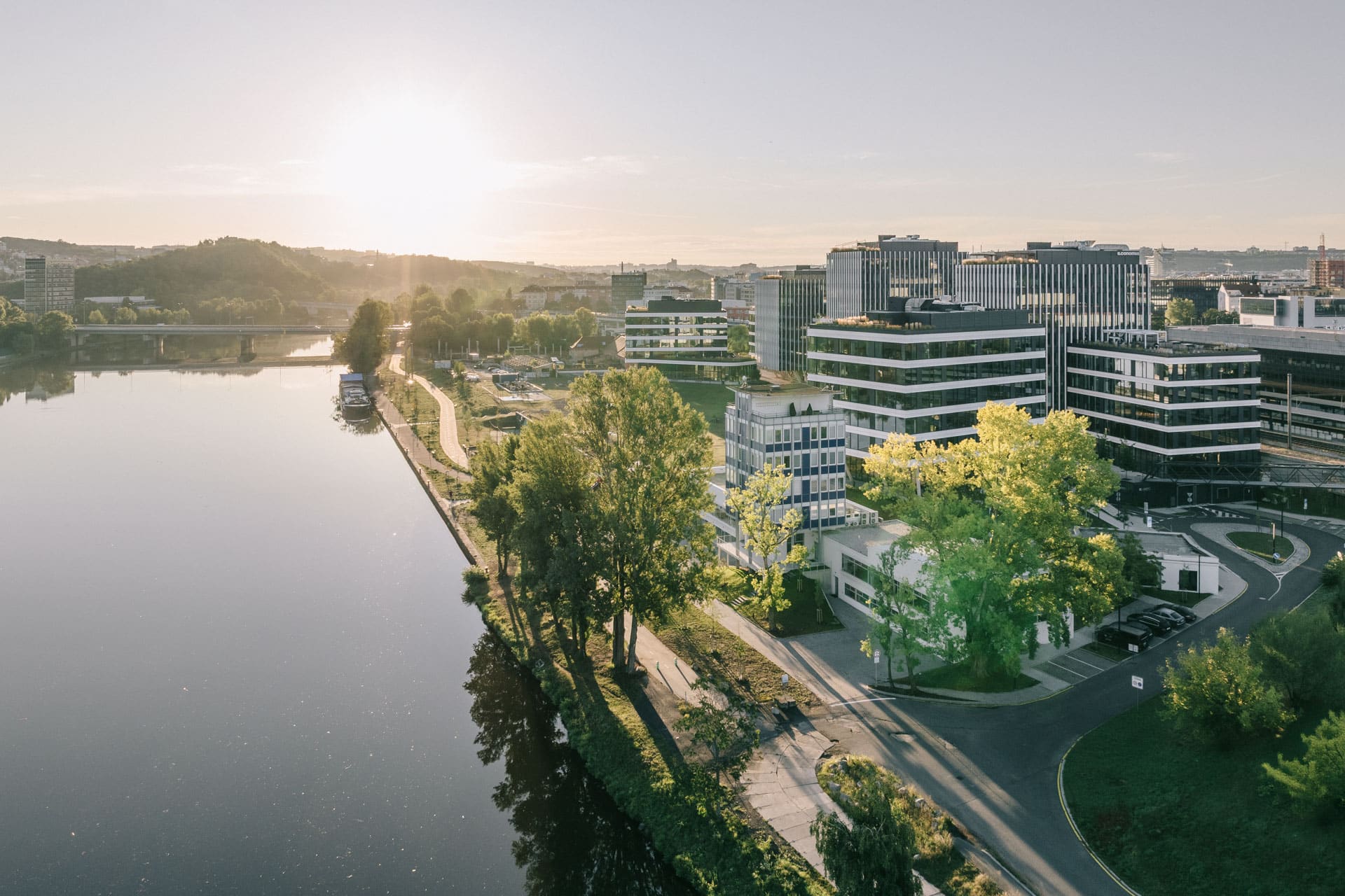 Město u řeky za východu slunce s moderními budovami a klidnou vodní hladinou. Stromy lemují cestu kolem řeky za krásného rána. Úžasná architektura a příroda.