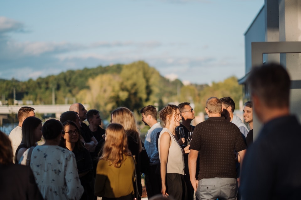 Skupina lidí na venkovní akci za slunečného dne se baví a směje v neformálním prostředí. V pozadí je vidět zelená krajina a modrá obloha s obláčky.