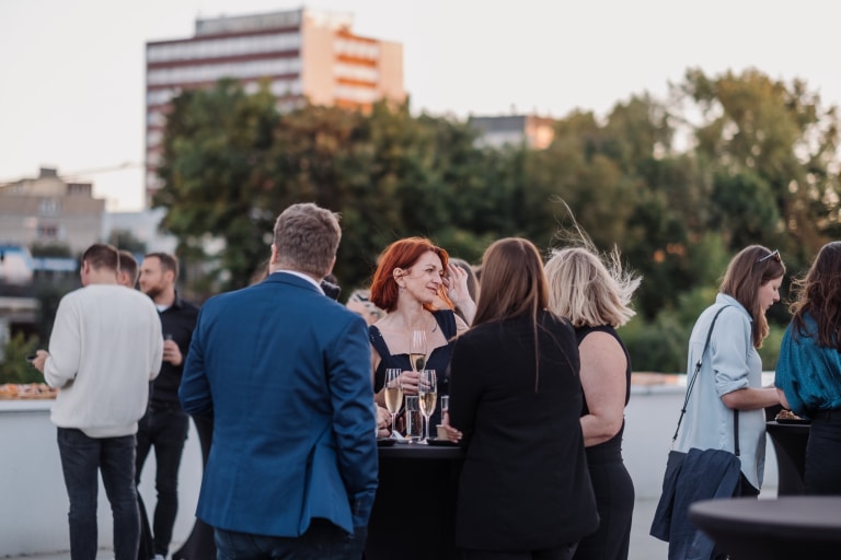 Lidé se scházejí na střešní terase pro společenskou akci za klidného večera v městské části obklopeni zelení a moderními budovami v pozadí.
